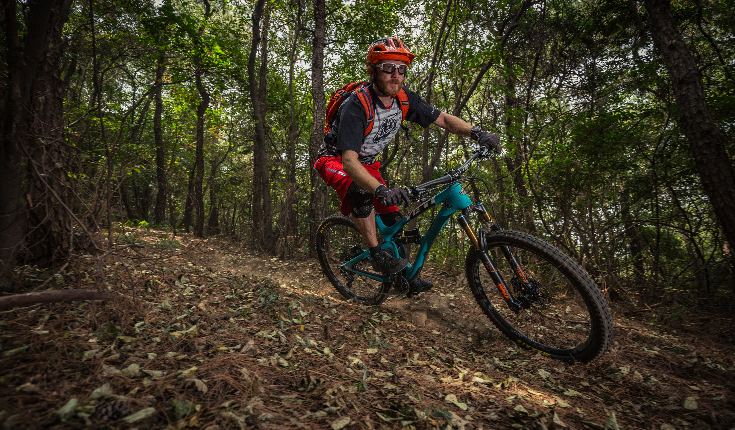 man riding a teal mountain bike through a forest with leaves